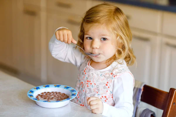 可爱的蹒跚学步的小女孩早餐吃着健康的麦片和牛奶。可爱的孩子，穿着五彩缤纷的衣服坐在厨房里，一边准备燕麦、谷类，一边玩乐。家里的门 — 图库照片