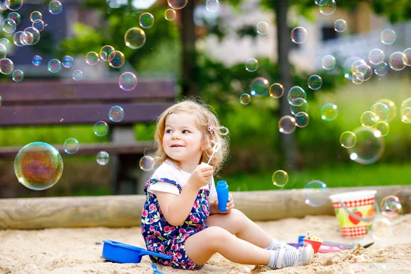 美しい小さなブロンドの幼児の女の子は、シャボン玉ブロワーを吹いて楽しんでいます。晴れた夏の日に遊び場で遊んでいるかわいいかわいい赤ちゃん。幸せなアクティブ面白い健康的な子供 — ストック写真