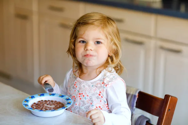 사랑 스러운 아장아장 걷는 소녀가 아침 식사로 우유와 함께 건강 한 시 리얼을 먹습니다. 다채 로운 옷을 입은 행복 한 아기를 부엌에 앉아서 귀리나 곡류 요리를 즐기도록 하라. 집안에 있는 집 — 스톡 사진