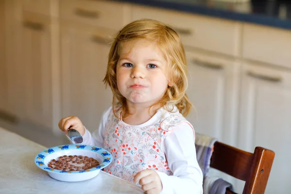 Menina adorável criança comendo cereais saudáveis com leite para o café da manhã. Bonito bebê feliz criança em roupas coloridas sentado na cozinha e se divertindo com a preparação de aveia, cereais. Interior em casa — Fotografia de Stock