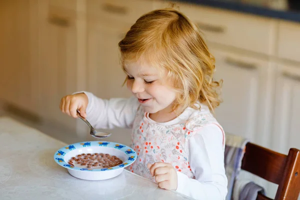 Menina adorável criança comendo cereais saudáveis com leite para o café da manhã. Bonito bebê feliz criança em roupas coloridas sentado na cozinha e se divertindo com a preparação de aveia, cereais. Interior em casa — Fotografia de Stock