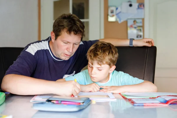 Mignon petit écolier garçon à la maison faire ses devoirs avec papa. Petit enfant écrivant avec des crayons colorés, père l'aidant, à l'intérieur. École primaire et éducation. — Photo