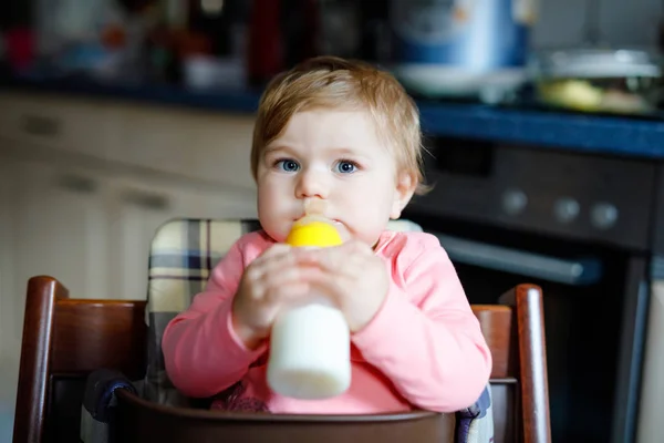 Linda menina adorável segurando mamadeira e bebendo leite fórmula. Primeira comida para bebés. Criança recém-nascida, sentada na cadeira da cozinha doméstica. Bebês saudáveis e conceito de alimentação por mamadeira — Fotografia de Stock