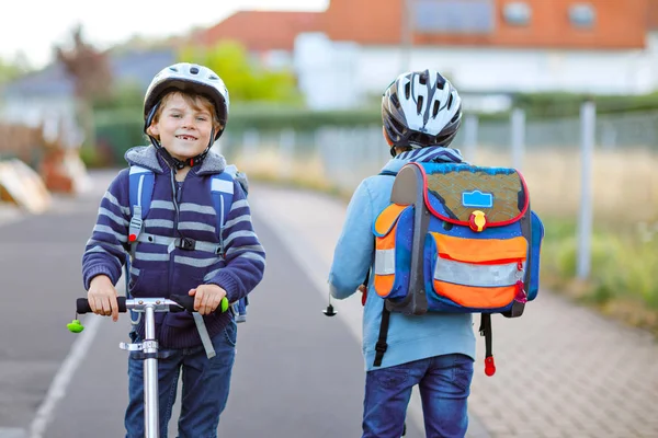 Zwei Schulkinder mit Schutzhelm fahren an einem sonnigen Tag mit dem Roller in der Stadt mit dem Rucksack. Fröhliche Kinder in bunten Kleidern radeln auf dem Schulweg. — Stockfoto