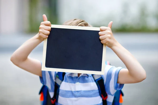 バックパックやカバンを持つ幸せな小さな子供の男の子。学校へ行く途中の小学生。健康な愛らしい子供屋外コピースペースのためのチョークデスク付き。学校や学校に戻る認識できない顔 — ストック写真