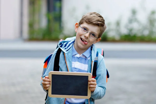 バックパックやランドセルとメガネを持つ幸せな小さな子供の男の子。学校へ行く途中の小学生。屋外で健康な愛らしい子供。手にコピースペースのための空のチョークデスク。学校の外または学校に戻る — ストック写真