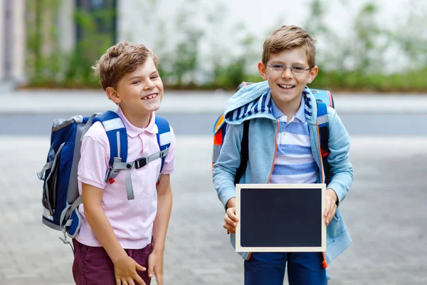 Två små pojkar med ryggsäck eller väska. Skolbarn på väg till skolan. Friska barn, bröder och bästa vänner utomhus tillbaka till skolan. Tomma krita skrivbord för copyspace. Lyckliga syskon. — Stockfoto