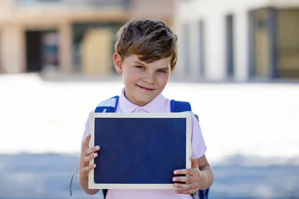 Niño feliz con mochila o mochila y gafas. Un colegial de camino a la escuela. Un niño sano y adorable al aire libre. Escritorio de tiza vacío para copiar espacio en las manos. Escuelas fuera o de vuelta a la escuela —  Fotos de Stock