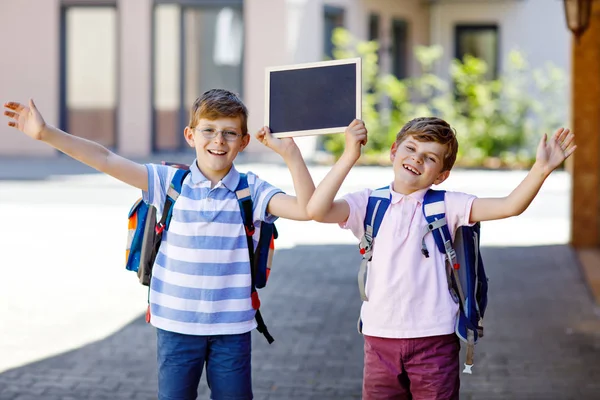 Två små pojkar med ryggsäck eller väska. Skolbarn på väg till skolan. Friska barn, bröder och bästa vänner utomhus tillbaka till skolan. Tomma krita skrivbord för copyspace. Lyckliga syskon. — Stockfoto