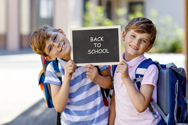 Dos niños pequeños con mochila o mochila. Los colegiales de camino a la escuela. Niños sanos, hermanos y mejores amigos al aire libre en la calle. De vuelta a la escuela en el escritorio de tiza. Felices hermanos. — Foto de Stock