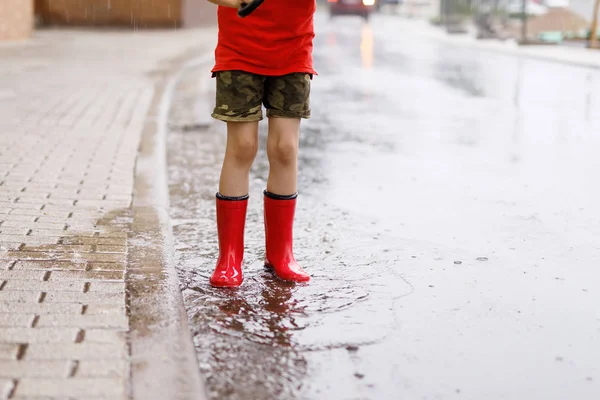 Kind met rode regenlaarzen die in een plas springen. Sluit maar af. Hij heeft plezier met spetteren met water. Warme zware zomerregen en blije kinderen — Stockfoto