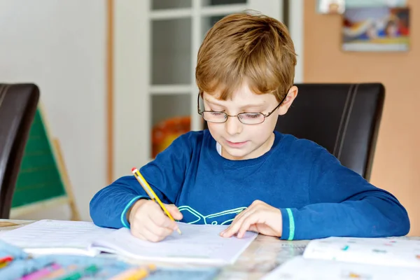 Portret van schattige school jongen jongen met bril thuis maken van huiswerk. Weinig geconcentreerd kind schrijven met kleurrijke potloden, binnenshuis. Basisschool en onderwijs — Stockfoto