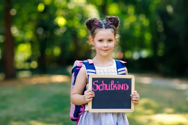 Niña feliz de pie con escritorio y mochila o mochila. Colegial en el primer día de clase elemental. Niño sano y adorable al aire libre, en el parque verde. Sobre el escritorio Colegial en alemán — Foto de Stock