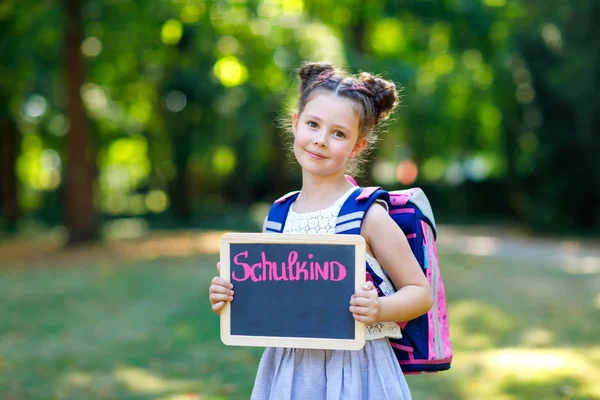 Fröhliches kleines Mädchen mit Schreibtisch und Rucksack oder Schulranzen. Schulkind am ersten Tag der Grundschule. gesundes liebenswertes Kind im Freien, im grünen Park. auf dem Schreibtisch - Schulkind — Stockfoto