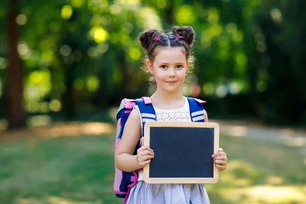 Счастливая маленькая девочка, стоящая со столом, рюкзаком или сумкой. Школьник в первый день начального класса. Здоровый очаровательный ребенок на открытом воздухе, в зеленом парке. Copyspace пустой стол — стоковое фото