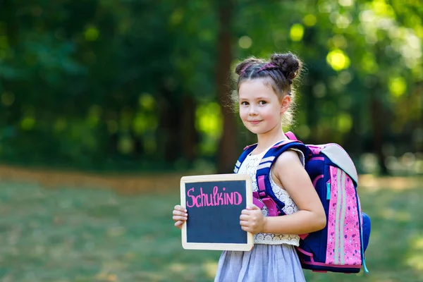Щасливий маленька дівчинка дитина стоїть з письмовим столом і рюкзаком або сумка. Школяр в перший день елементарного класу. Здорова чарівна дитина на відкритому повітрі, в зеленому парку. На столі-шкільна дитина-на німецькій — стокове фото