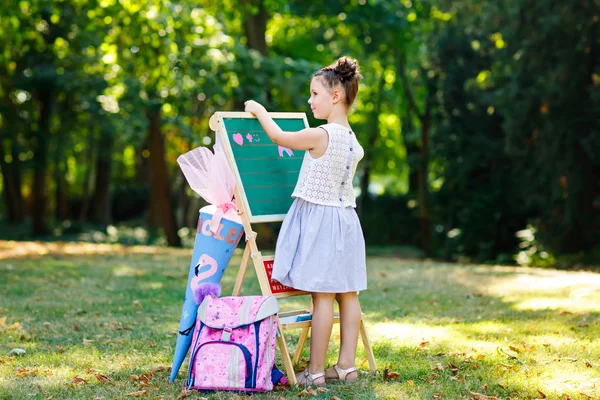 Счастливая маленькая девочка стоит у большого стола с мелом. школьник в первый день начального класса с сумкой и традиционным подарочным конусом. Здоровый ребенок пишет и рисует на открытом воздухе. Копировальное пространство на столе. — стоковое фото