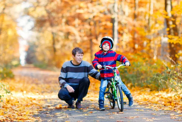 Liten förskola kid pojke och hans far i höst park med en cykel. Pappa undervisning sonen cykling. Aktiv familj fritid. Barn med hjälm på cykel. Säkerhet, sport, fritid med barnen koncept — Stockfoto