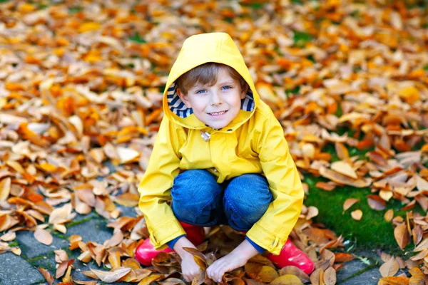 Portrait of happy cute little kid boy in yellow rain coat and red rubber boots with autumn leaves background. Funny child having fun and playing in fall forest or park on cold autumnal day — Stock Photo, Image