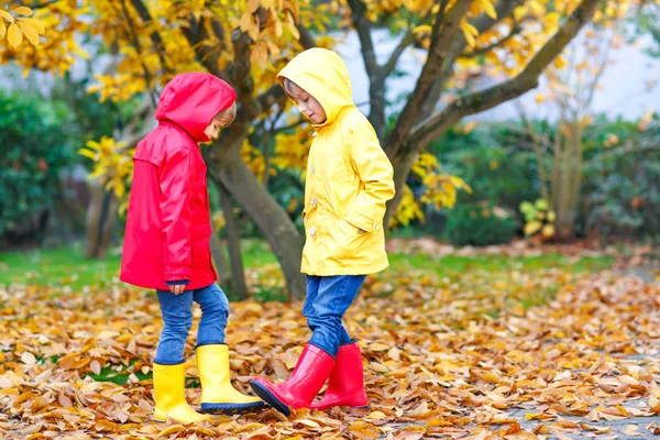 Two little best friends and kids boys autumn park in colorful clothes. Happy siblings children having fun in red and yellow rain coats and rubber boots. Family playing outdoors. active leisure. — Stock Photo, Image