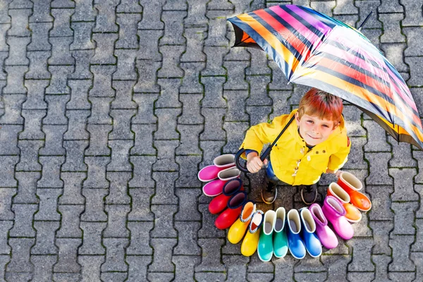 Gyerek kisfiú és a csoport színes eső csizma. Szőke gyermek állandó égisze alatt. Közeli kép a schoolkid és a különböző gumi csizma, a magas, szög. Esős őszi cipő — Stock Fotó