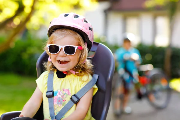 Retrato de una niña pequeña con casco de seguridad en la cabeza sentada en el asiento de la bicicleta de los padres. Niño en bicicleta en el fondo. Concepto de protección segura e infantil. Viaje familiar y de fin de semana. — Foto de Stock