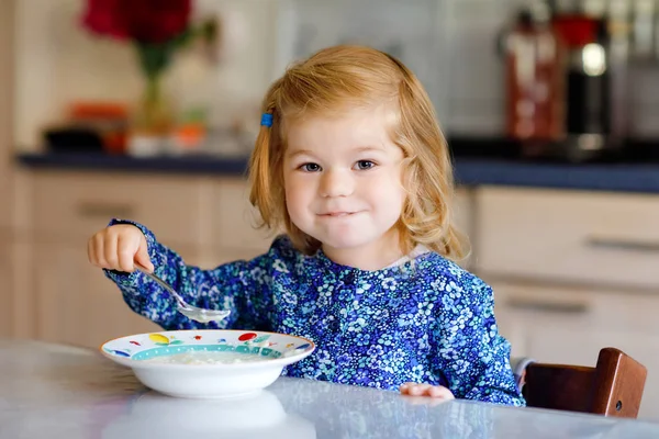 おいしい幼児の女の子は朝食のためにミルクと健康的なシリアルを食べています。キッチンに座って、オート、シリアルを準備するのが楽しいカラフルな服のかわいい幸せな赤ちゃん。家の中で — ストック写真