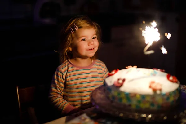 Entzückendes kleines Mädchen, das seinen zweiten Geburtstag feiert. Baby Kind isst Marshmellows Dekoration auf hausgemachtem Kuchen, drinnen. Glückliches gesundes Kleinkind überrascht über Feuerwerkskörper auf Kuchen — Stockfoto