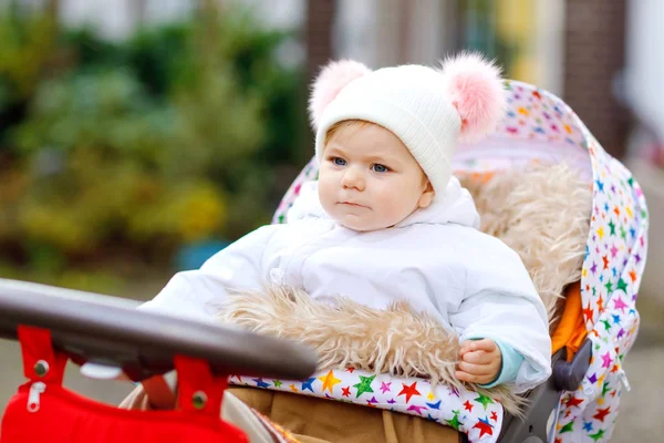 Schattige kleine mooie baby meisje zit in de kinderwagen of wandelwagen op de herfst dag. Gelukkig gezond kind gaan voor een wandeling op frisse lucht in warme kleren. Baby met in kleurrijke kleding en hoed met bobbels — Stockfoto