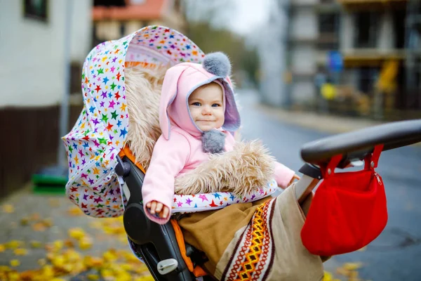 Bonito pequena menina bonita sentada no carrinho de bebê ou carrinho no dia de outono. Criança sorridente feliz em roupas quentes, moda elegante casaco de bebê rosa com orelhas de coelho. Bebê indo em uma caminhada com os pais. — Fotografia de Stock