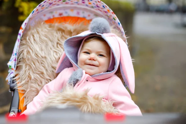 Cute little beautiful baby girl sitting in the pram or stroller on autumn day. Happy smiling child in warm clothes, fashion stylish pink baby coat with bunny ears. Baby going on a walk with parents. — Stock Photo, Image