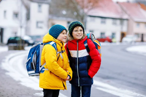 Zwei kleine Jungen der Grundschulklasse laufen bei Schneefall zur Schule. Glückliche Kinder, die Spaß haben und mit dem ersten Schnee spielen. Geschwister und beste Freunde mit Rucksack in bunter Winterkleidung. — Stockfoto