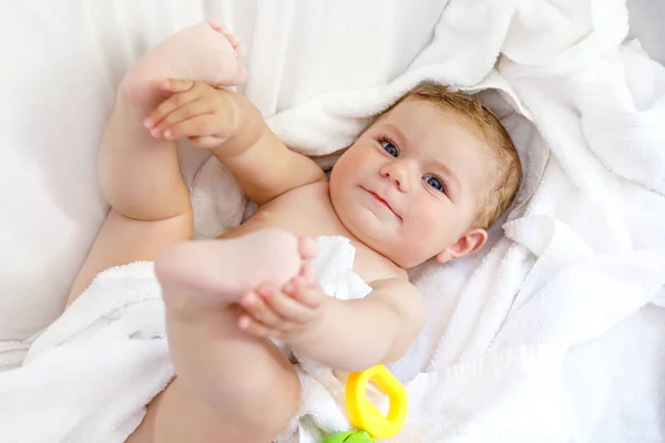 Χαριτωμένο μικρό μωρό παίζει με παιχνίδι κουδουνίστρα και δικά της πόδια μετά το ανάληψη μπάνιο. Αξιολάτρευτο όμορφο κορίτσι τυλιγμένο σε λευκές πετσέτες — Φωτογραφία Αρχείου