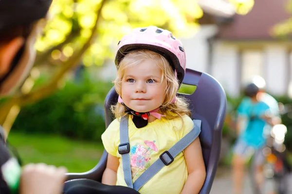 Portrait of little toddler girl with security helmet on the head sitting in bike seat of parents. Boy on bicycle on background. Safe and child protection concept. Family and weekend activity trip.