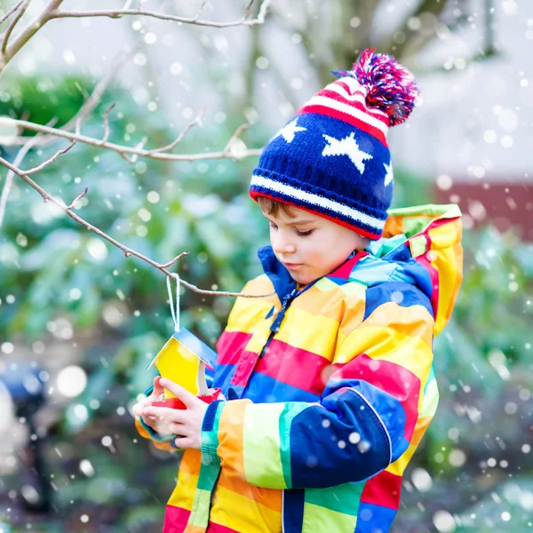 Niño pequeño alimentando pájaros en invierno. Lindo niño preescolar feliz colgando colorido casa de aves hechas a sí mismo en el árbol en el frío día helado. Niño en ropa wam colorido poniendo semillas en alimentador . —  Fotos de Stock