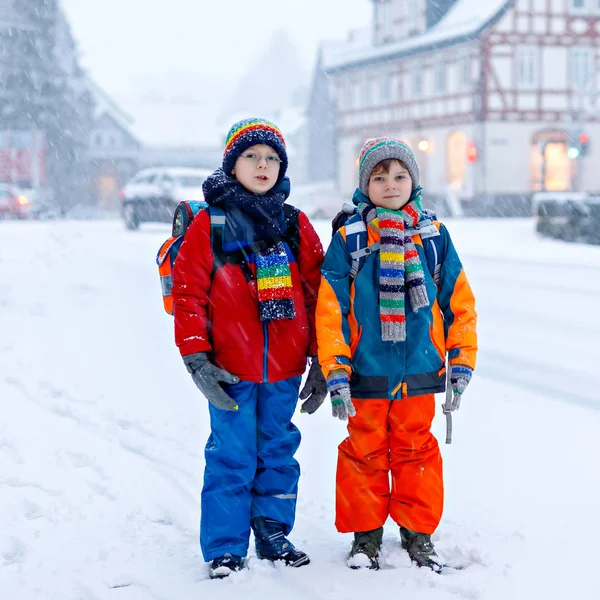 Zwei kleine Jungen der Grundschulklasse laufen bei Schneefall zur Schule. Glückliche Kinder, die Spaß haben und mit dem ersten Schnee spielen. Geschwister und Freunde mit Rucksack in bunter Winterkleidung. — Stockfoto