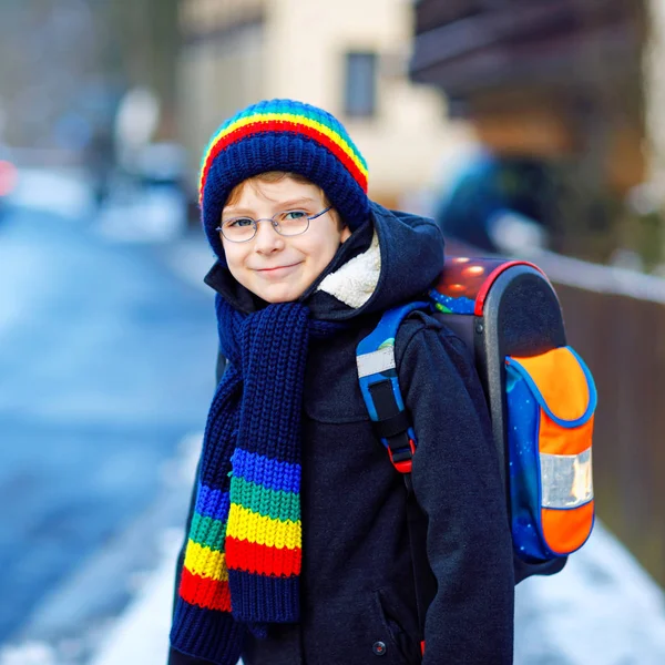 Chłopiec z podstawówki, idący do szkoły podczas opadów śniegu. Szczęśliwe zdrowe dziecko w okularach zabawy i zabawy ze śniegiem. Z plecakiem lub torbą w kolorowe zimowe ubrania. — Zdjęcie stockowe