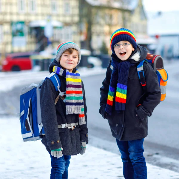 Dwójka małych chłopców z podstawówki, idących do szkoły podczas opadów śniegu. Szczęśliwe dzieci bawiące się i bawiące z pierwszym śniegiem. Rodzeństwo przyjaciele z plecakiem w kolorowe ubrania zimowe. — Zdjęcie stockowe