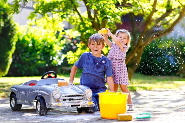 Двое счастливых детей моют большую старую игрушечную машину в летнем саду — стоковое фото