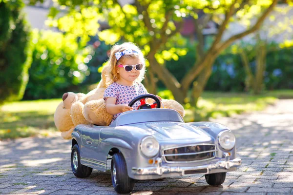 Little adorable toddler girl driving big vintage toy car and having fun with playing with plush toy bear, outdoors. Gorgeous happy healthy child enjoying warm summer day. Smiling stunning kid in gaden — Stock Photo, Image
