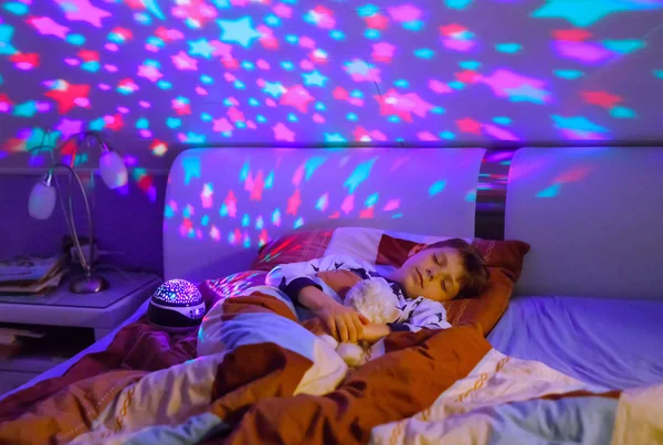 Menino dormindo na cama com lâmpada colorida. Criança da escola sonhando e segurando brinquedo de pelúcia. Miúdo zangado com a escuridão. Luz noturna mudando cores, estrelas e lua e tocando música. — Fotografia de Stock