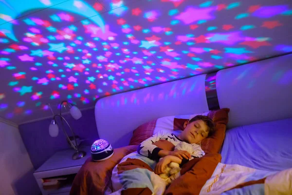 Niño durmiendo en la cama con una lámpara de colores. Niño de la escuela soñando y sosteniendo peluche. Un chico enojado por la oscuridad. Luz nocturna cambiando colores, estrellas y luna y tocando música. — Foto de Stock