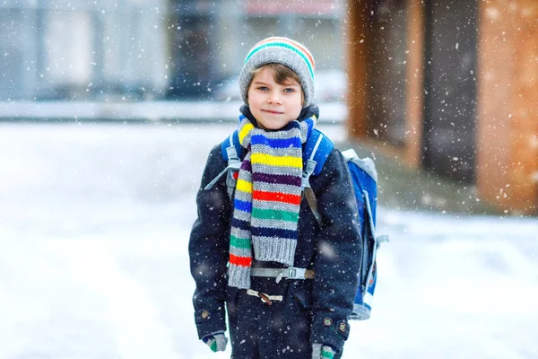 Kleine schooljongen van de lagere klasse die naar school loopt tijdens de sneeuwval. Gelukkig kind dat plezier heeft en speelt met de eerste sneeuw. Leerling met rugzak of tas in kleurrijke winterkleding. — Stockfoto