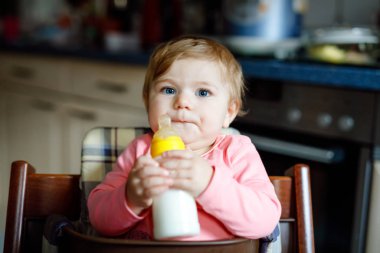 Sevimli, tapılası, süt şişesi tutan ve mama sütü içen kız bebek. Bebekler için ilk yemek. Yeni doğmuş çocuk, evcil mutfağın sandalyesinde oturuyor. Sağlıklı bebekler ve şişe besleme konsepti