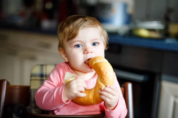 Bonita menina bebê comendo pão. Criança adorável comendo pela primeira vez pedaço de pretzel ou croissant. Criança feliz e sorridente saudável. Os primeiros dentes vêm aí. Em casa dentro de casa na cozinha . — Fotografia de Stock