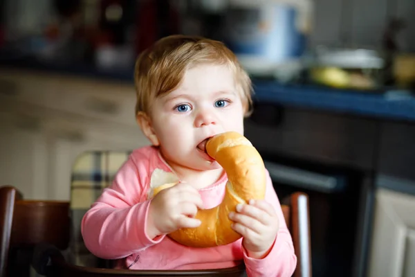 Linda niñita comiendo pan. Adorable niño comiendo por primera vez pedazo de pretzel o croissant. Niño feliz sonriente saludable. Vienen los primeros dientes. En casa dentro de la cocina . — Foto de Stock