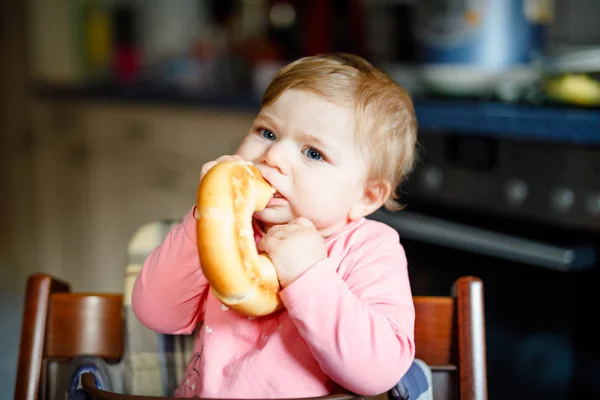 Linda niñita comiendo pan. Adorable niño comiendo por primera vez pedazo de pretzel o croissant. Niño feliz sonriente saludable. Vienen los primeros dientes. En casa dentro de la cocina . —  Fotos de Stock
