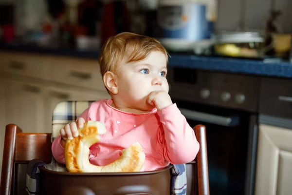 Bonita menina bebê comendo pão. Criança adorável comendo pela primeira vez pedaço de pretzel ou croissant. Criança feliz e sorridente saudável. Os primeiros dentes vêm aí. Em casa dentro de casa na cozinha . — Fotografia de Stock