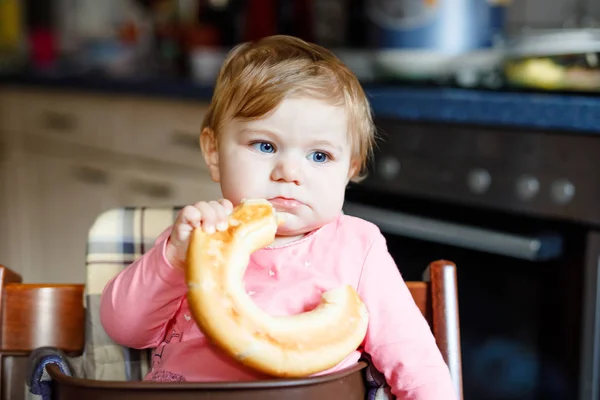 Cute little baby girl eating bread. Adorable child eating for the first time piece of pretzel or croissant. Healthy smiling happy child. First teeth coming. At home indoors in the kitchen. — Stock Photo, Image