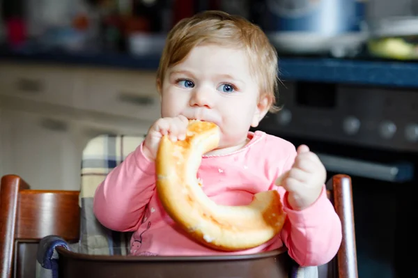 Cute little baby girl eating bread. Adorable child eating for the first time piece of pretzel or croissant. Healthy smiling happy child. First teeth coming. At home indoors in the kitchen. — Stock Photo, Image